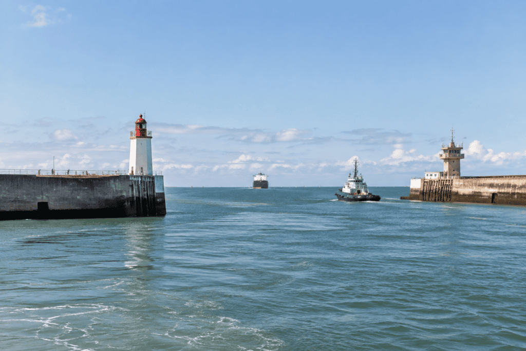 Un remorqueur avance dans le chenal entre deux jetées, près d’un phare et d’une tour de signalisation.