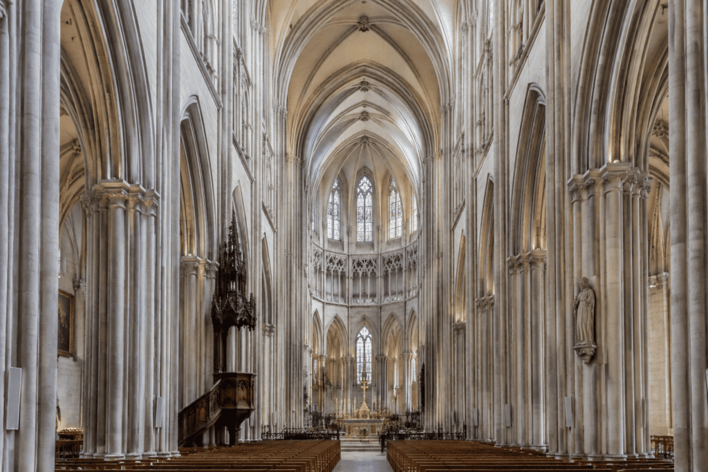 Nef gothique avec de hautes colonnes, des voûtes nervurées, des vitraux et des bancs.