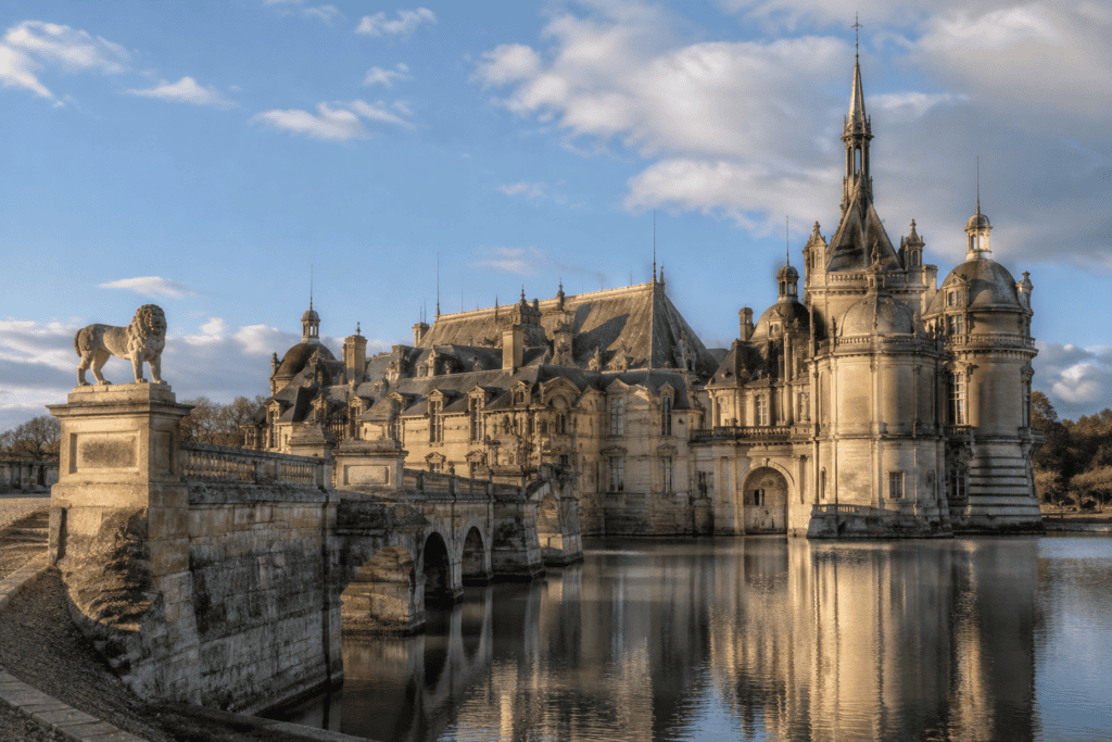 Le Château de Chantilly se reflète dans l’eau, derrière un pont de pierre orné d’une statue de lion.