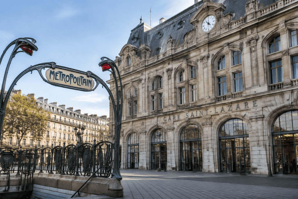Façade de la gare Saint-Lazare avec son horloge et une entrée de métro Guimard au premier plan.