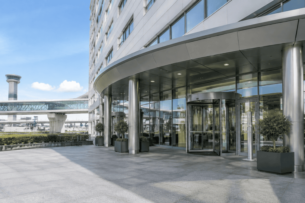 Entrée vitrée sous un auvent incurvé, devant un bâtiment moderne, avec une passerelle surélevée à proximité.