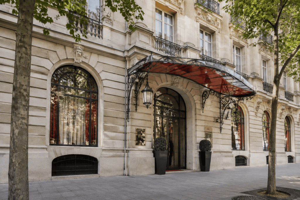 Façade en pierre claire avec une entrée sous un auvent rouge et noir, encadrée d’arbres.