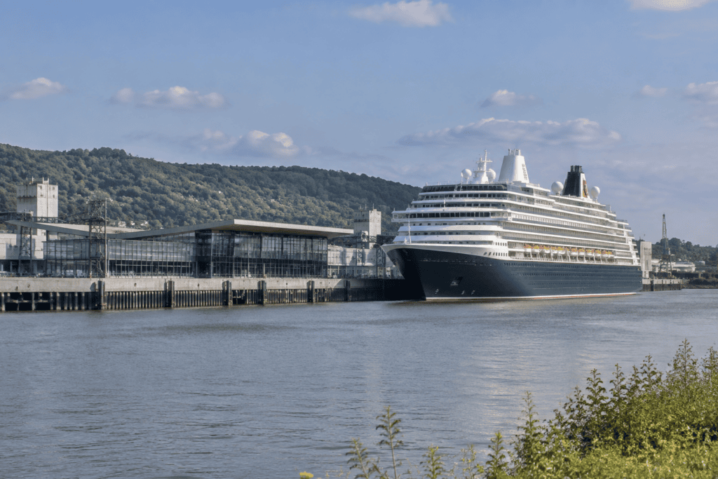 Un grand navire de croisière est amarré devant des bâtiments portuaires, de l’autre côté du fleuve.