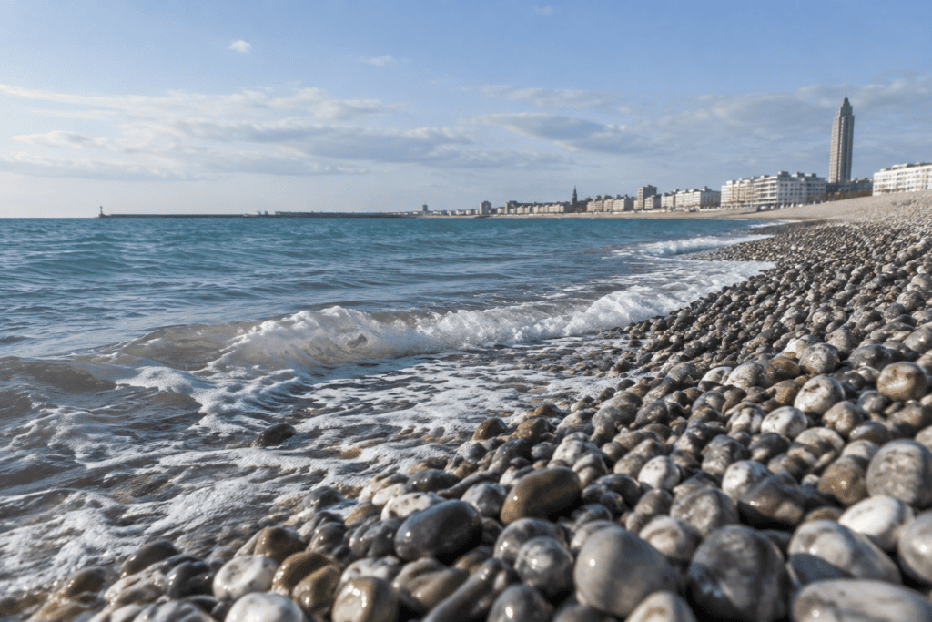 Des galets au premier plan bordent les vagues de la Manche, avec la ville du Havre visible le long du rivage au loin.