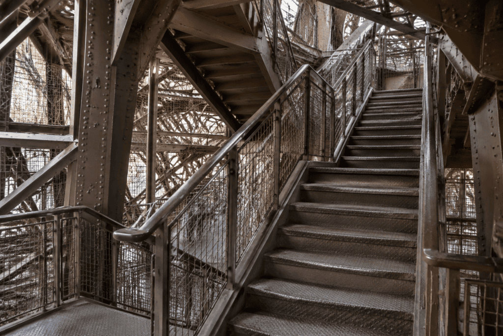 Marches métalliques et garde-corps d’un escalier intérieur, entourés de poutrelles rivetées.