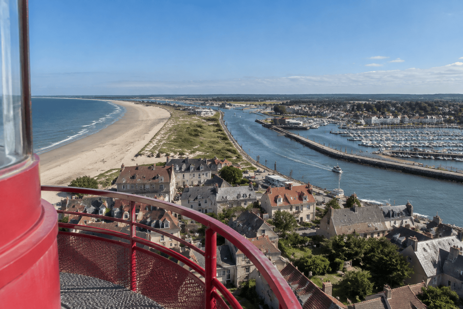 Vue en hauteur sur une plage, des maisons, un chenal et un port de plaisance, avec une rambarde rouge au premier plan.