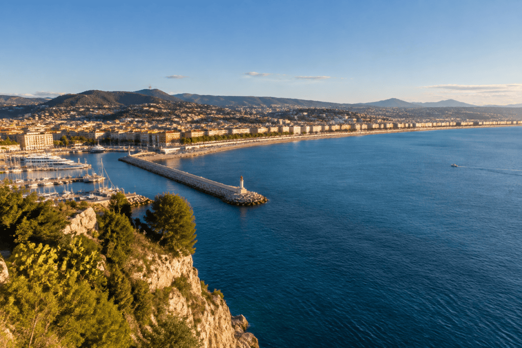 Vue en hauteur sur le port de Nice, la baie et le littoral bordé d’immeubles, avec des collines à l’horizon.