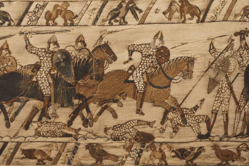 Détail brodé montrant des cavaliers casqués, des chevaux et des combattants, encadrés par des bordures figurées.