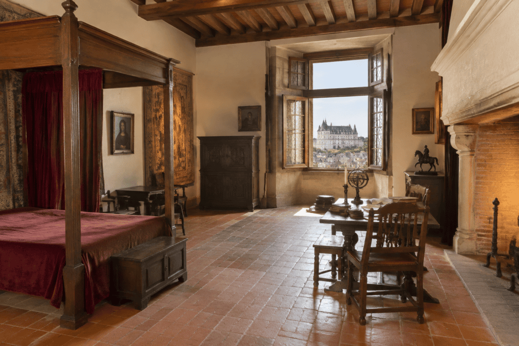 Chambre meublée avec un lit à baldaquin, une cheminée et une fenêtre ouverte sur un château et les toits d’une ville.