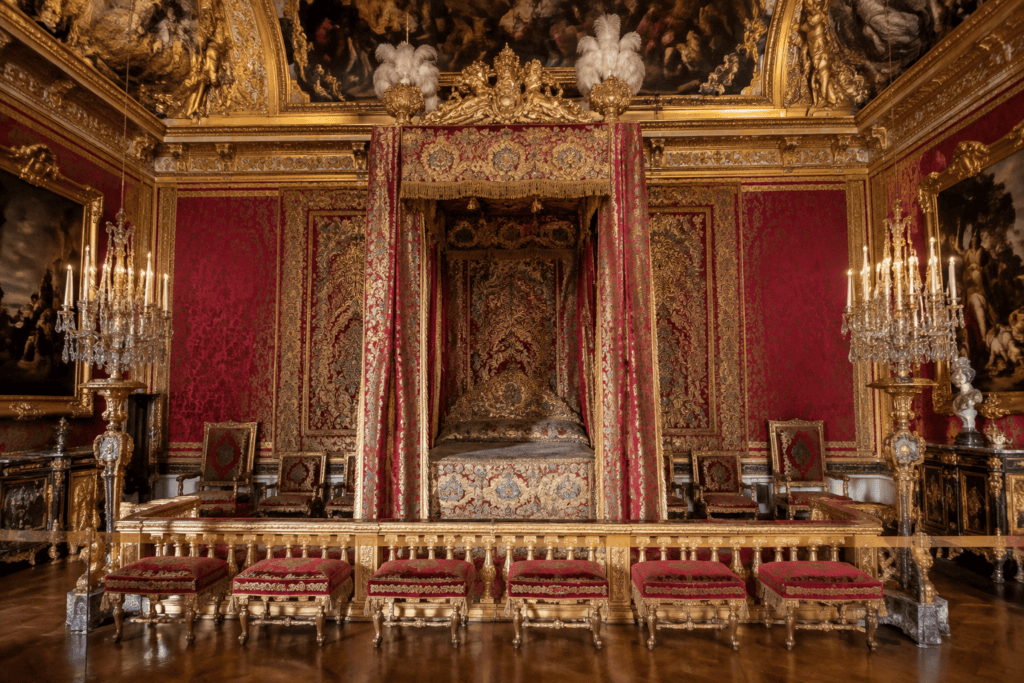 Lit d’apparat sous un baldaquin brodé, encadré de tentures rouges, de sièges et de chandeliers dans une chambre richement décorée.