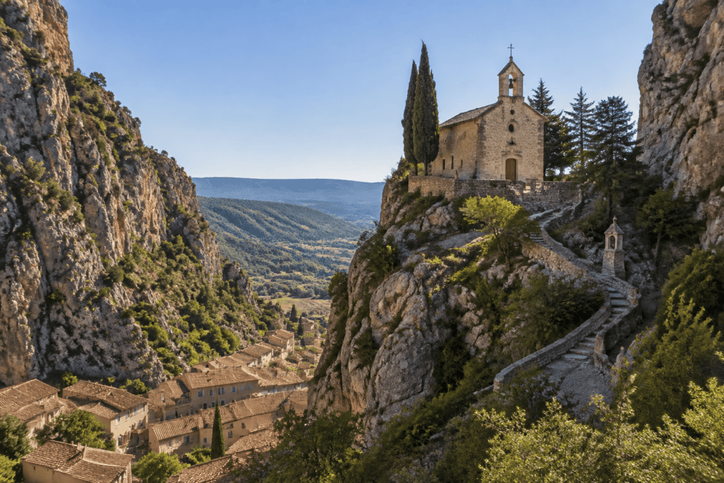 Une chapelle en pierre se dresse sur un promontoire rocheux, au-dessus des toits du village et entre des falaises calcaires ; un chemin en lacets y mo