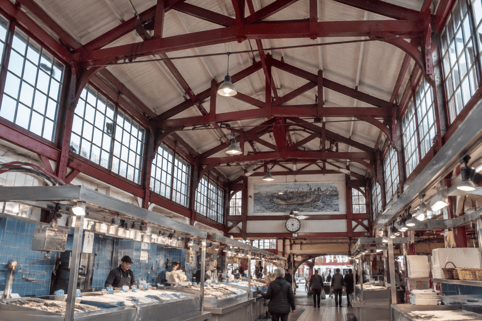 Des étals de poissons sous une charpente métallique rouge et de hautes fenêtres dans une halle de marché