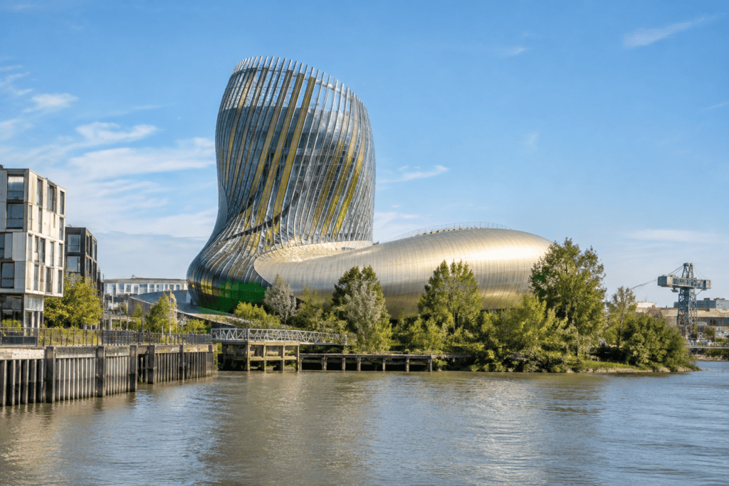 Le bâtiment incurvé de la Cité du Vin se dresse au bord de l’eau, entouré de végétation et de constructions urbaines.