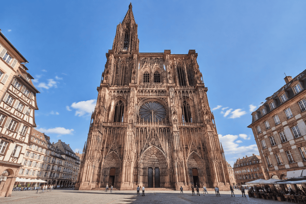 La façade gothique de la cathédrale Notre-Dame de Strasbourg, avec sa grande rosace et sa flèche, domine une place bordée de bâtiments.