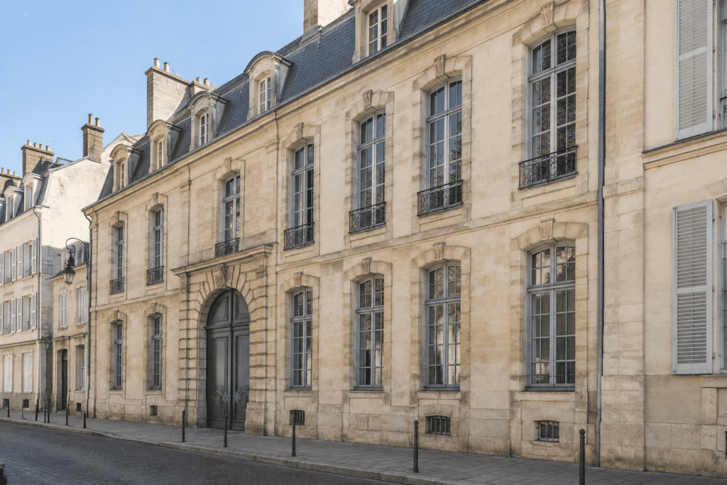 Façade en pierre claire d’un hôtel particulier, avec de hautes fenêtres à petits carreaux, des lucarnes et une porte cochère en arc.