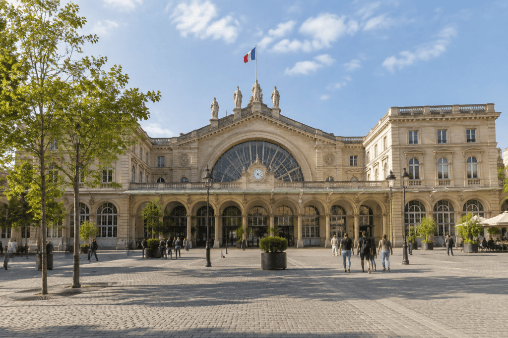 Façade en pierre de la Gare de l’Est, avec une grande verrière en arc, une horloge et des statues au-dessus de l’entrée.