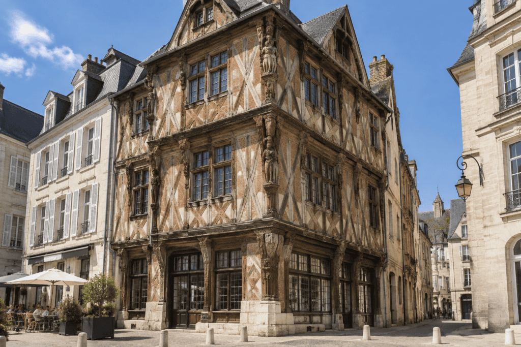 Maison ancienne à pans de bois, avec des figures sculptées sur l’angle et des façades donnant sur une place pavée.