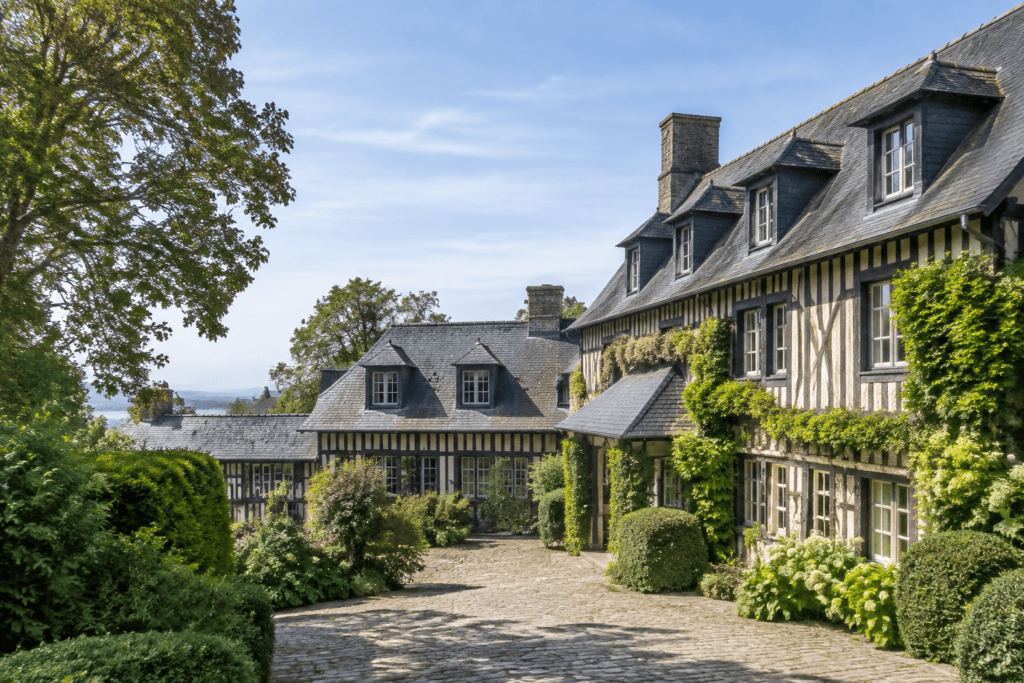 Façades à colombages et toitures en ardoise entourant une cour pavée, avec des arbustes et un grand arbre.