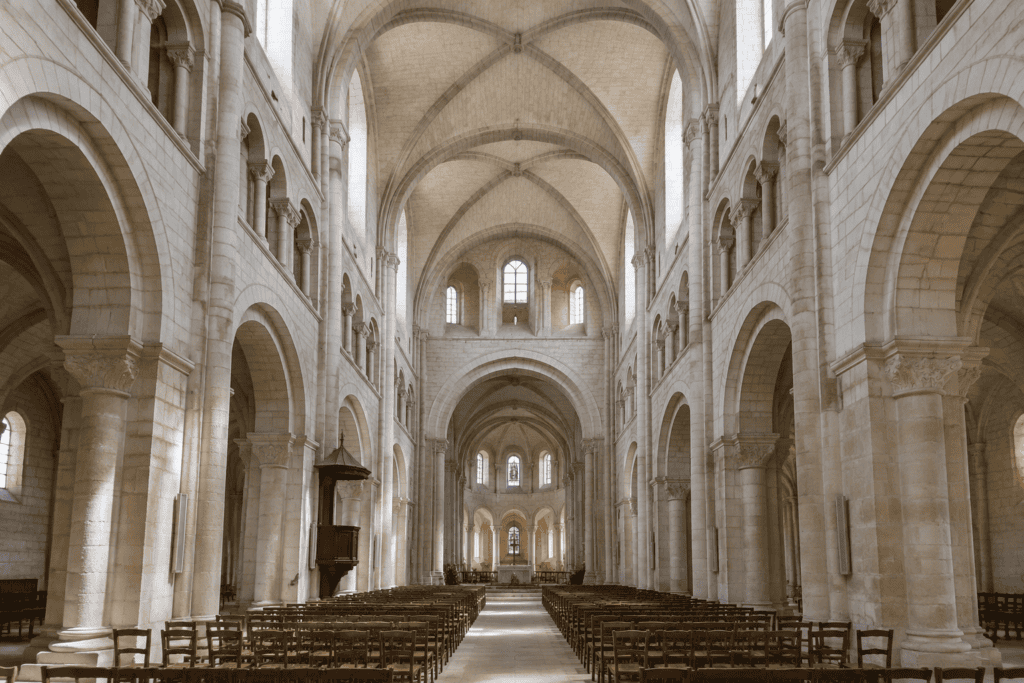 Nef d’une église romane, avec de hautes arcades, des voûtes croisées, des fenêtres hautes et des rangées de chaises.