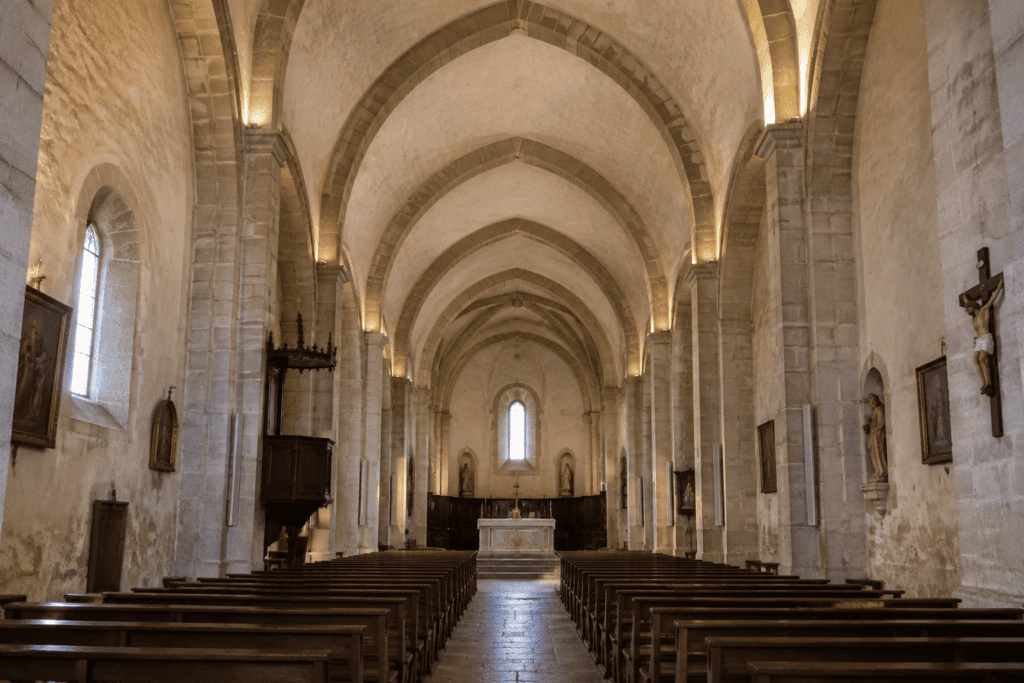 Vue axiale d’une nef d’église aux travées voûtées, avec des rangées de bancs, un autel au fond et des pilastres le long des murs.