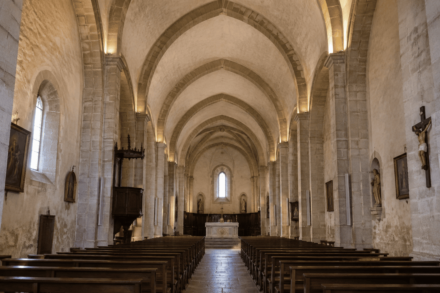 Vue axiale d’une nef d’église aux travées voûtées, avec des rangées de bancs, un autel au fond et des pilastres le long des murs.