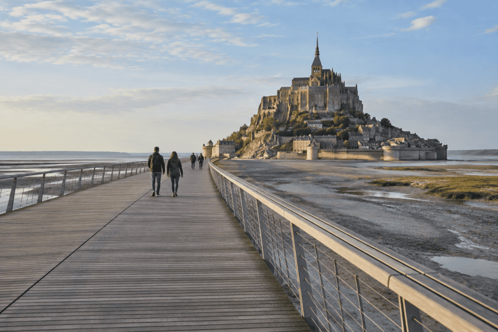 La passerelle traverse les grèves vers le Mont-Saint-Michel, visible au loin, avec quelques marcheurs.