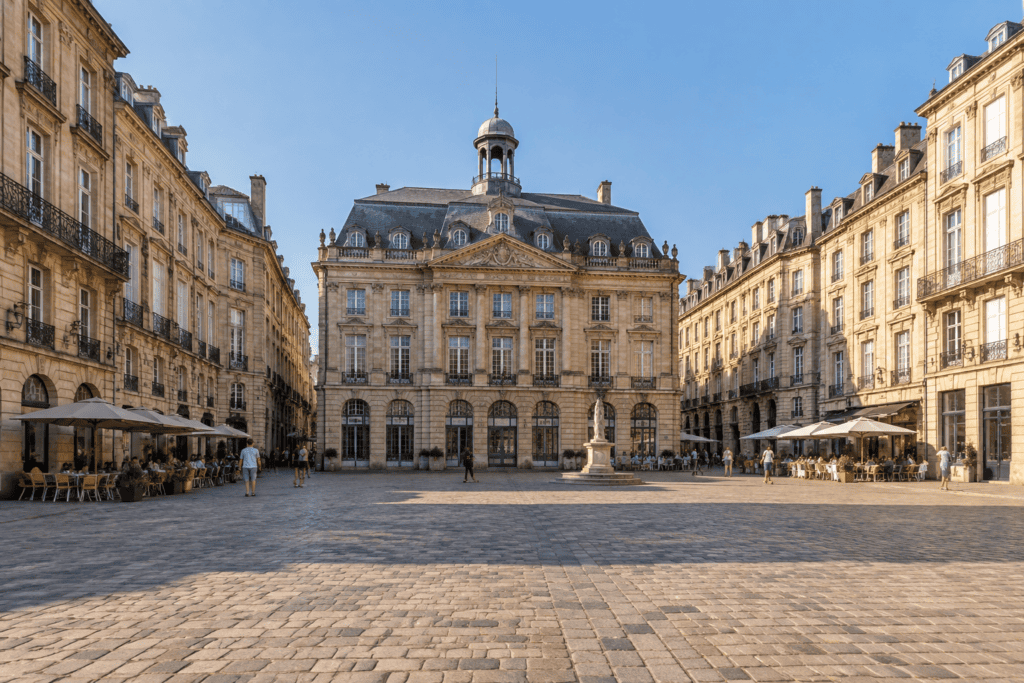 Place pavée entourée de façades en pierre et de terrasses de café, avec un bâtiment orné au fond.