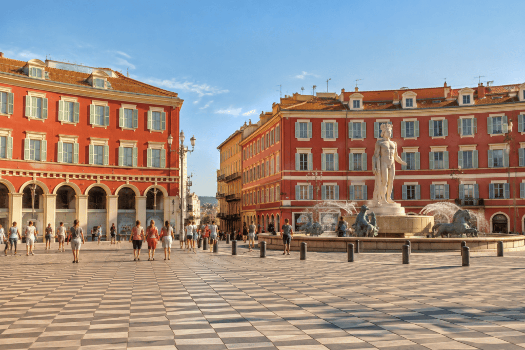 La place Masséna, avec ses façades rouges à arcades, son dallage à motifs et une fontaine ornée d’une statue.