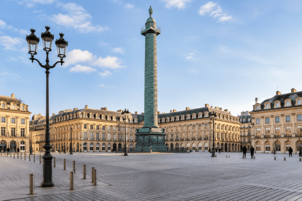 La colonne Vendôme au centre d’une place pavée, entourée de façades classiques et de quelques passants.