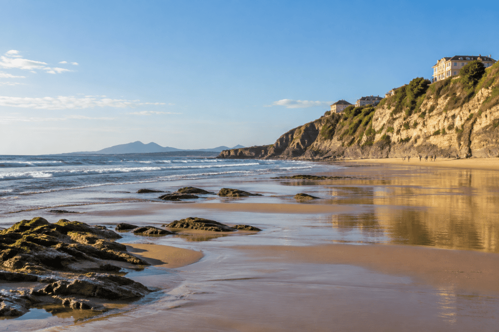 Une large plage découverte à marée basse s’étend au pied de falaises dorées, avec des rochers sur le sable et l’océan à gauche.