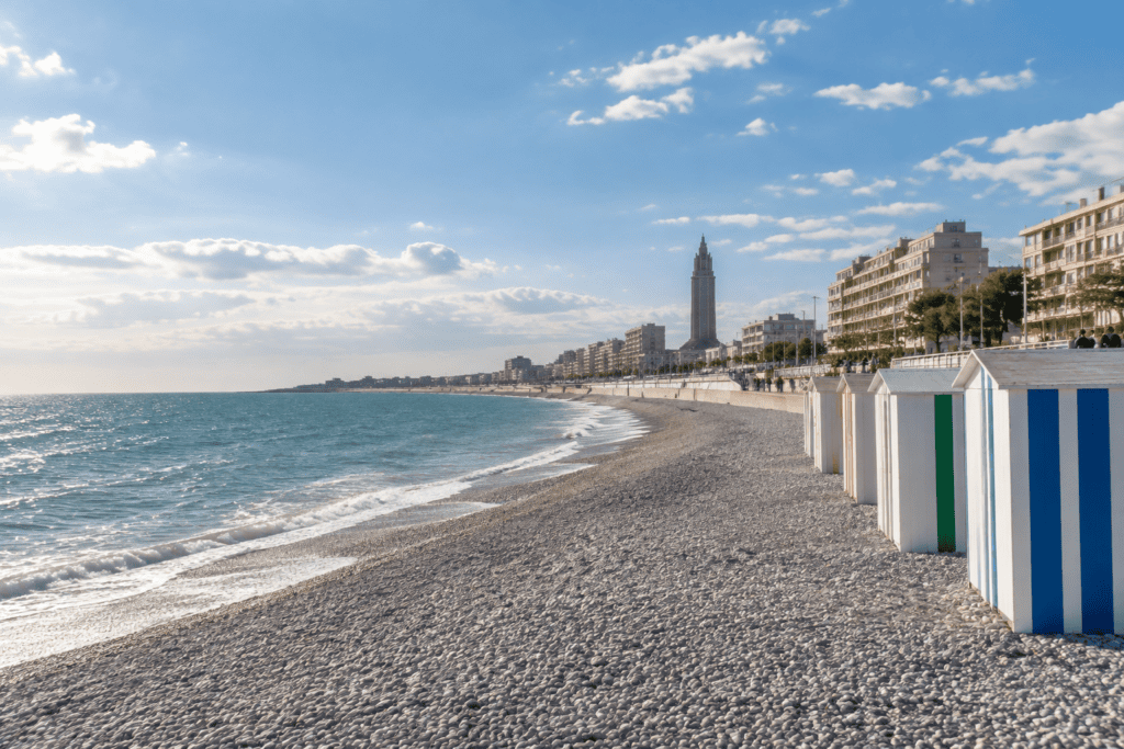 Des cabines de plage bordent une plage de galets face à la mer, avec les immeubles du front de mer en arrière-plan.