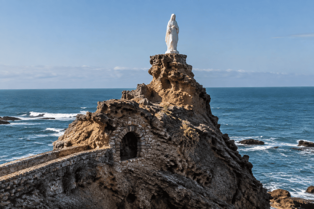 Une statue blanche se dresse sur un éperon rocheux au-dessus de l’Atlantique ; une ouverture voûtée apparaît dans la roche.