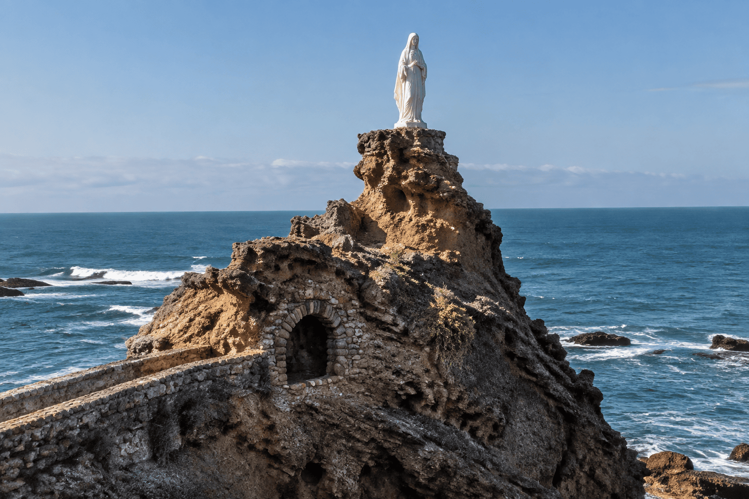 Une statue blanche se dresse sur un éperon rocheux au-dessus de l’Atlantique ; une ouverture voûtée apparaît dans la roche.