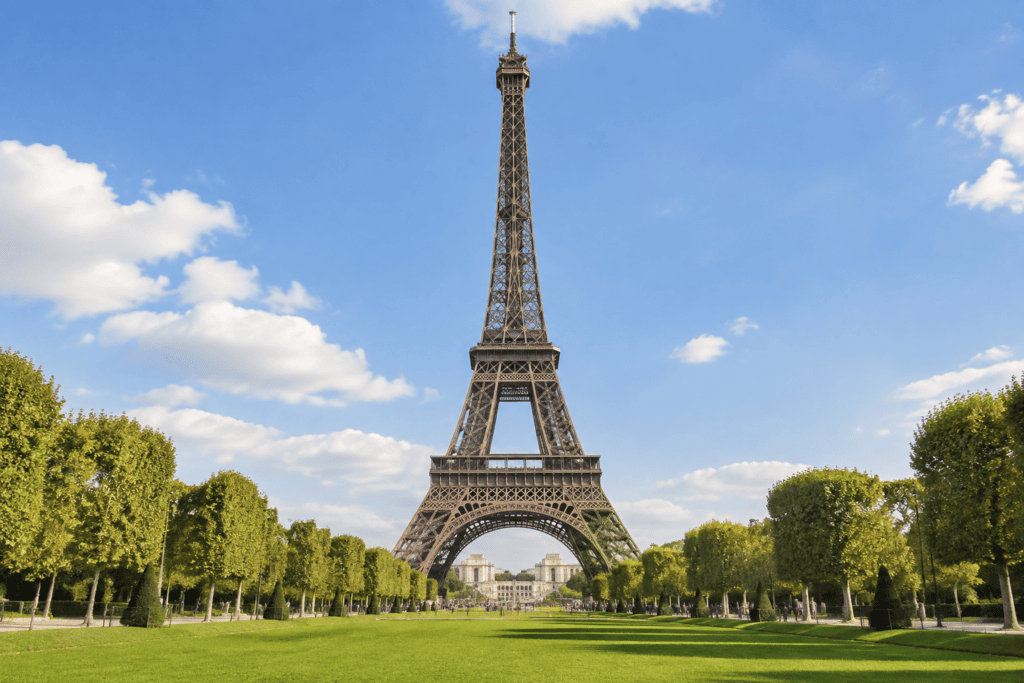 La tour Eiffel se dresse au fond d’une large pelouse bordée d’arbres, sous un ciel bleu.