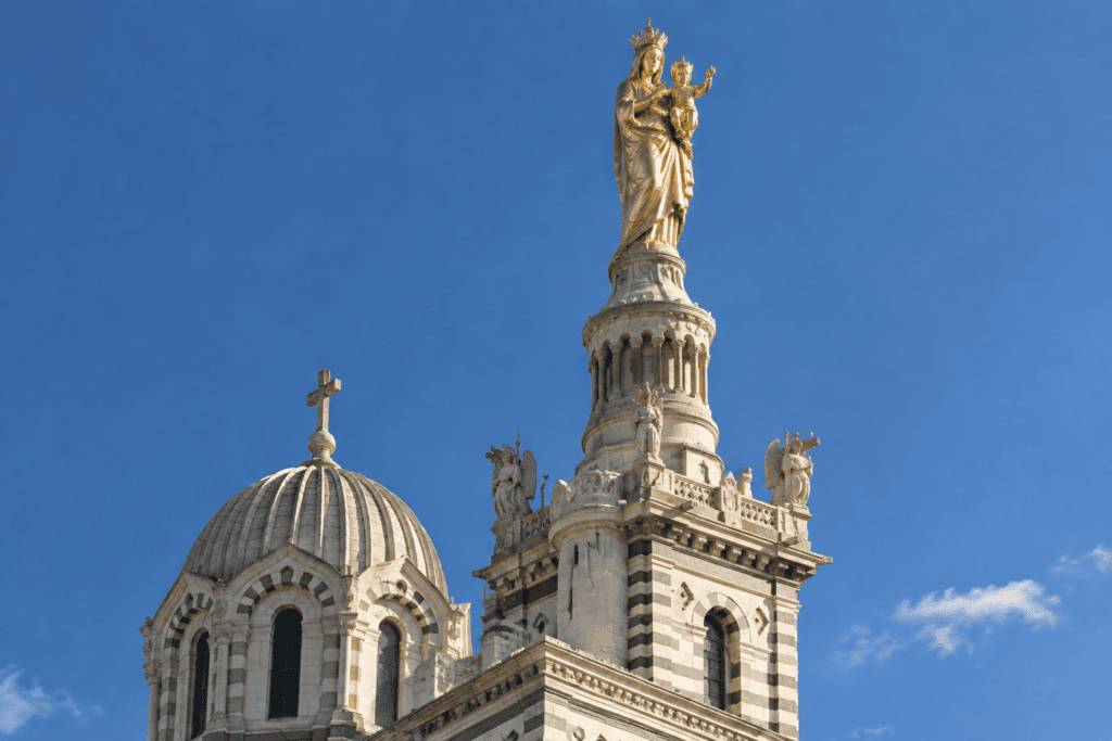 La statue dorée de la Vierge à l’Enfant couronne une tour de Notre-Dame de la Garde, à côté d’une coupole de pierre.