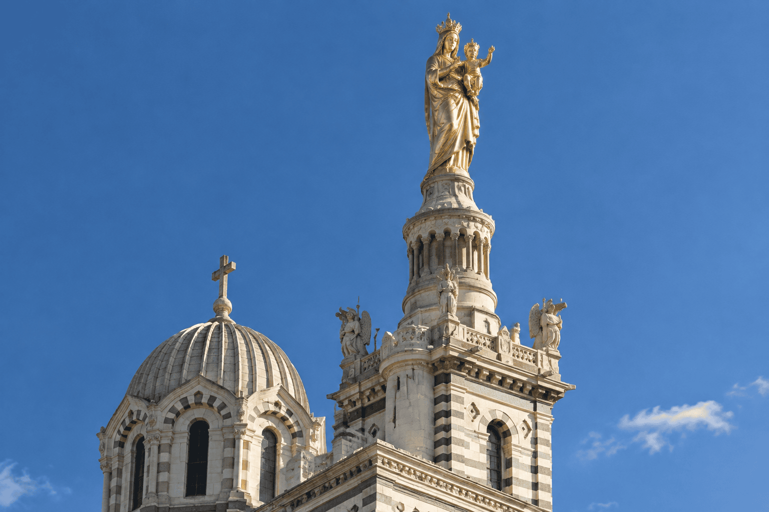 La statue dorée de la Vierge à l’Enfant couronne une tour de Notre-Dame de la Garde, à côté d’une coupole de pierre.