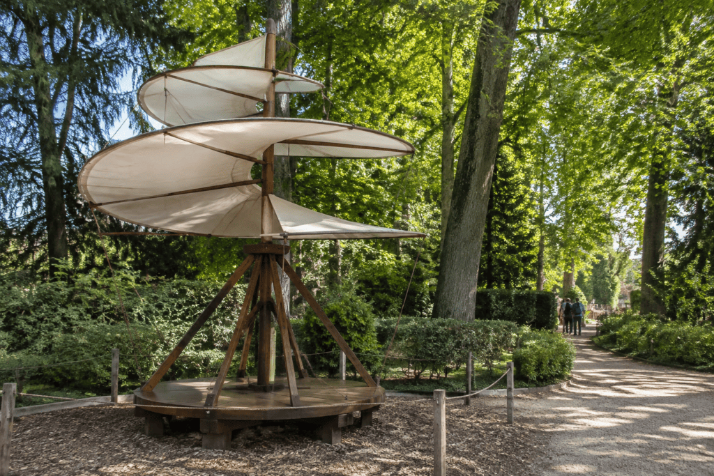Grande maquette à pales hélicoïdales installée parmi les arbres, au bord d’une allée.