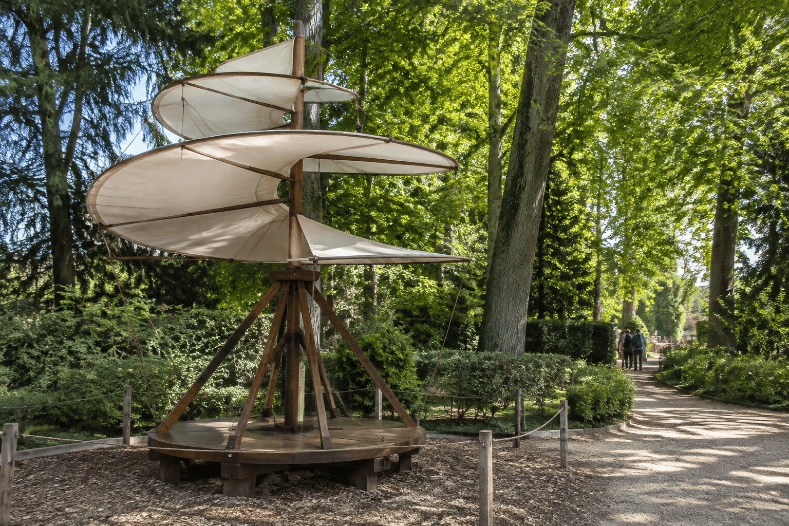 Grande maquette à pales hélicoïdales installée parmi les arbres, au bord d’une allée.
