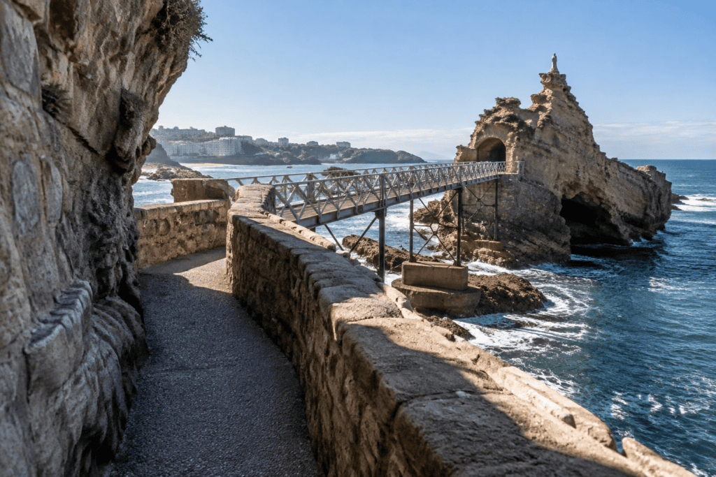 Un passage piétonnier longe une paroi et un muret de pierre avant de rejoindre la passerelle métallique menant au Rocher de la Vierge, au-dessus de l’