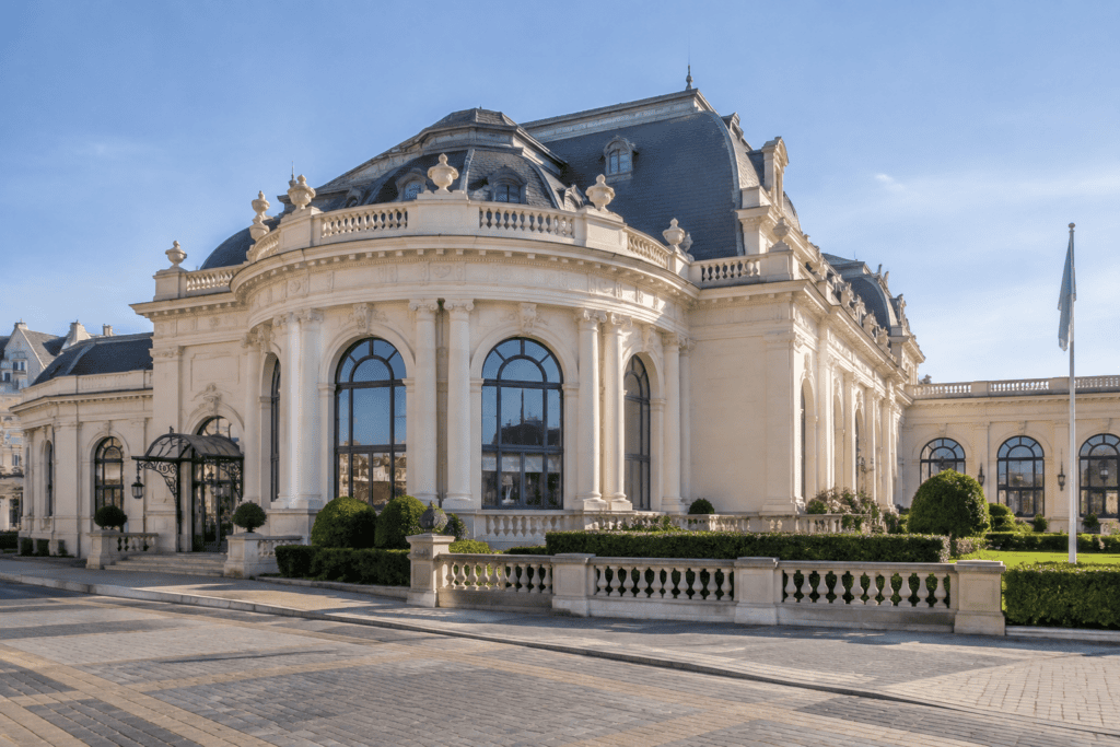 Façade du Casino Barrière Deauville Façade claire aux grandes fenêtres cintrées et au toit en ardoise, entourée de jardins taillés.