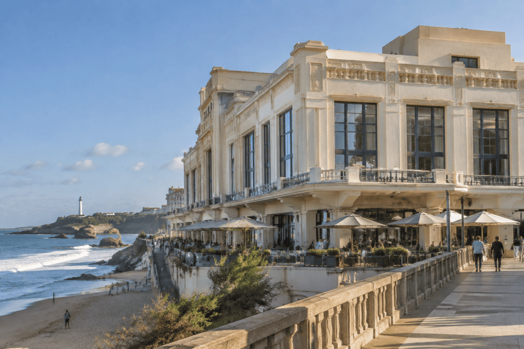 Façade claire du Casino de Biarritz et terrasse bordant la promenade, avec la plage et le phare visibles à gauche.