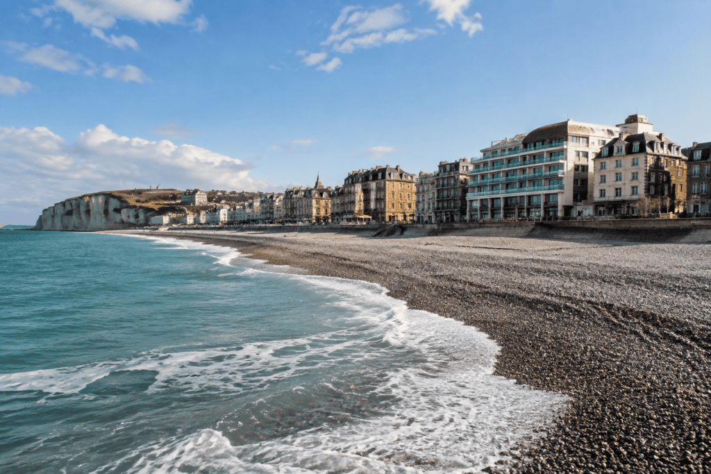 La mer borde une plage de galets devant une rangée de bâtiments du front de mer, sous un ciel dégagé.