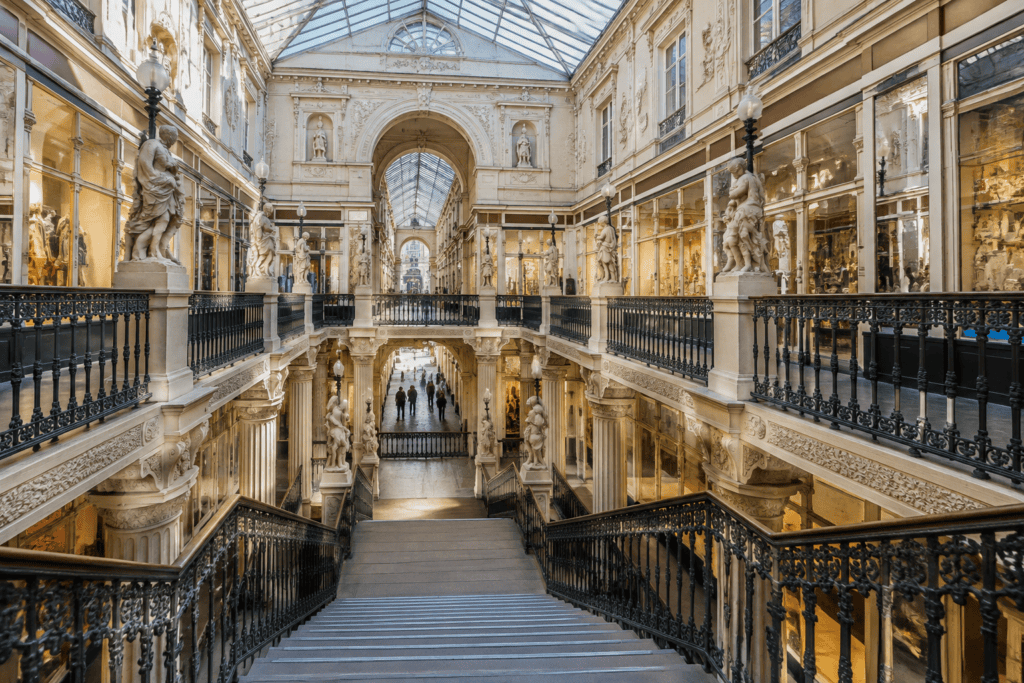 Grand escalier bordé de balustrades en fer forgé dans une galerie commerçante à plusieurs niveaux, sous une verrière.
