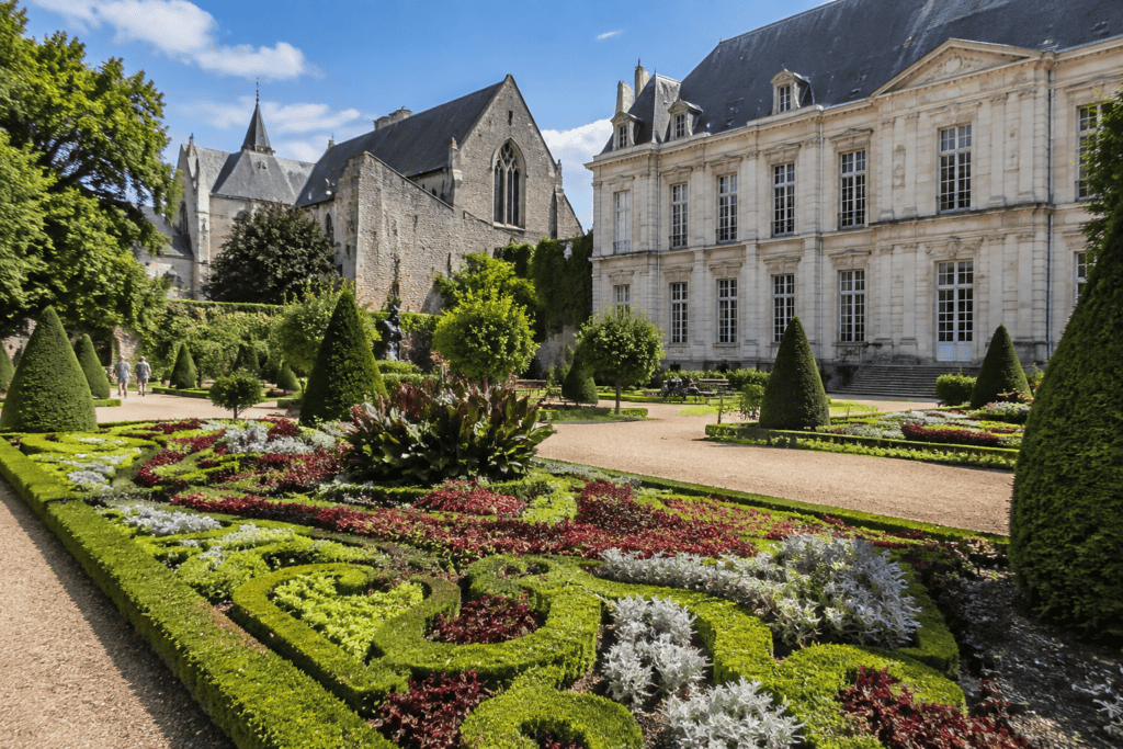 Parterres de fleurs et arbustes taillés dans un jardin formel, devant un grand bâtiment en pierre et une église gothique.