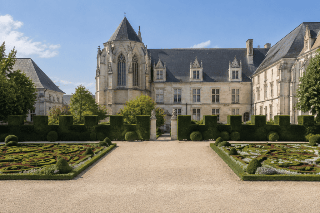 Deux parterres fleuris encadrent une large allée gravillonnée bordée d’une haie taillée, devant un palais ancien.