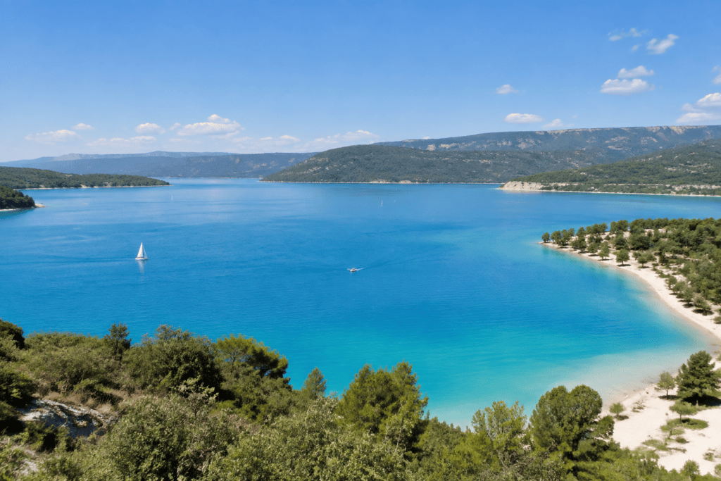 Une vaste étendue d’eau bleue s’étire entre des collines boisées et des rives claires ; un voilier navigue au loin.