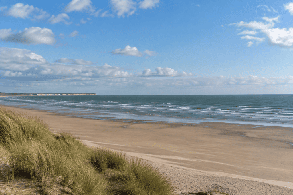 Plage de sable bordée de dunes herbeuses, face à la mer sous un ciel nuageux.