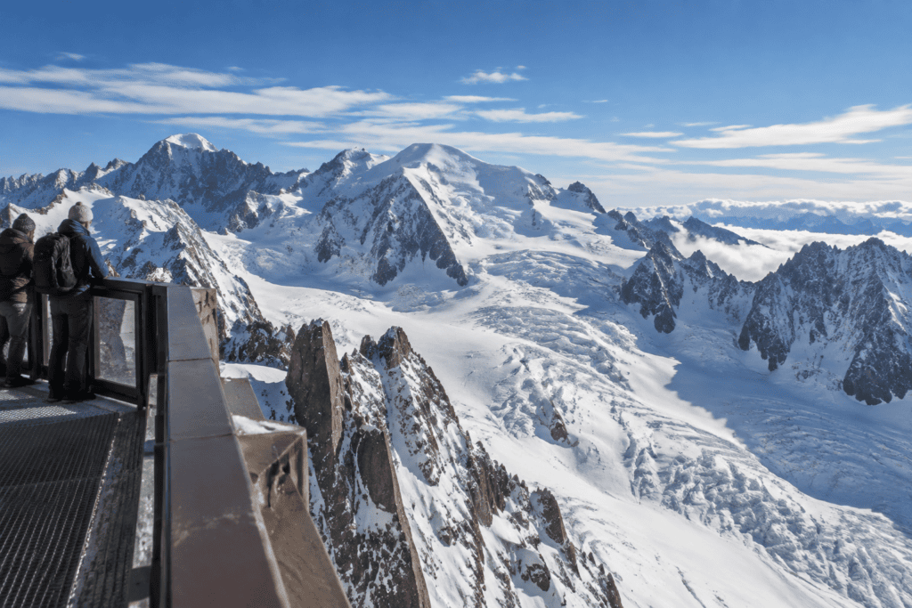 Des visiteurs regardent un vaste panorama de sommets enneigés et de glaciers depuis une terrasse avec garde-corps.