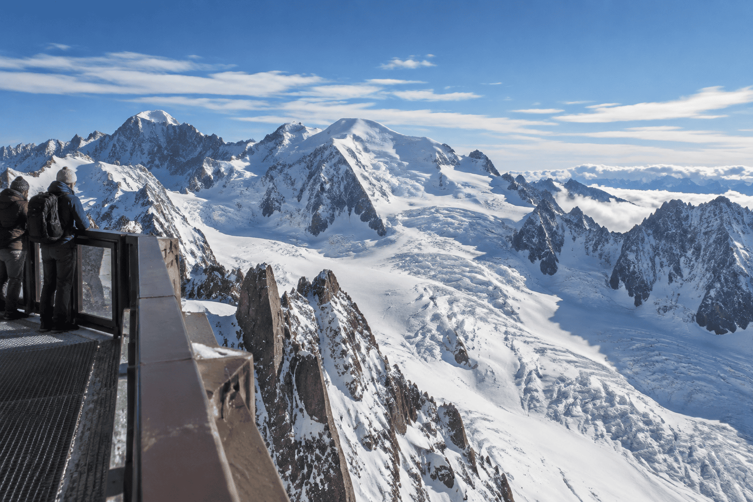 Des visiteurs regardent un vaste panorama de sommets enneigés et de glaciers depuis une terrasse avec garde-corps.