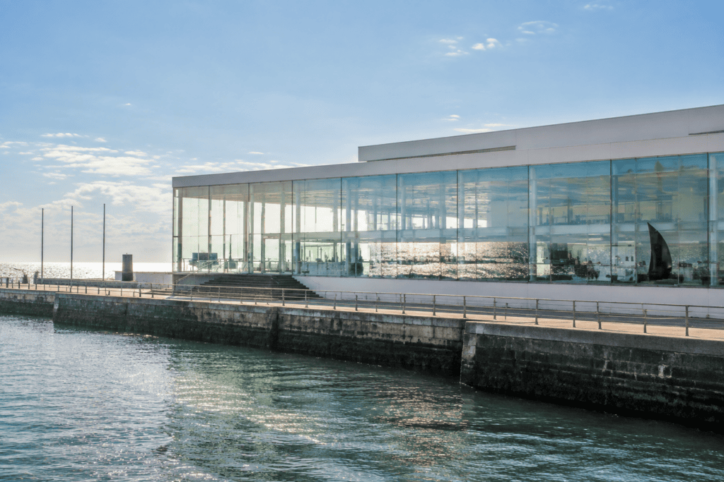 Façade vitrée d’un bâtiment contemporain au bord de l’eau, avec une promenade et un quai au premier plan.